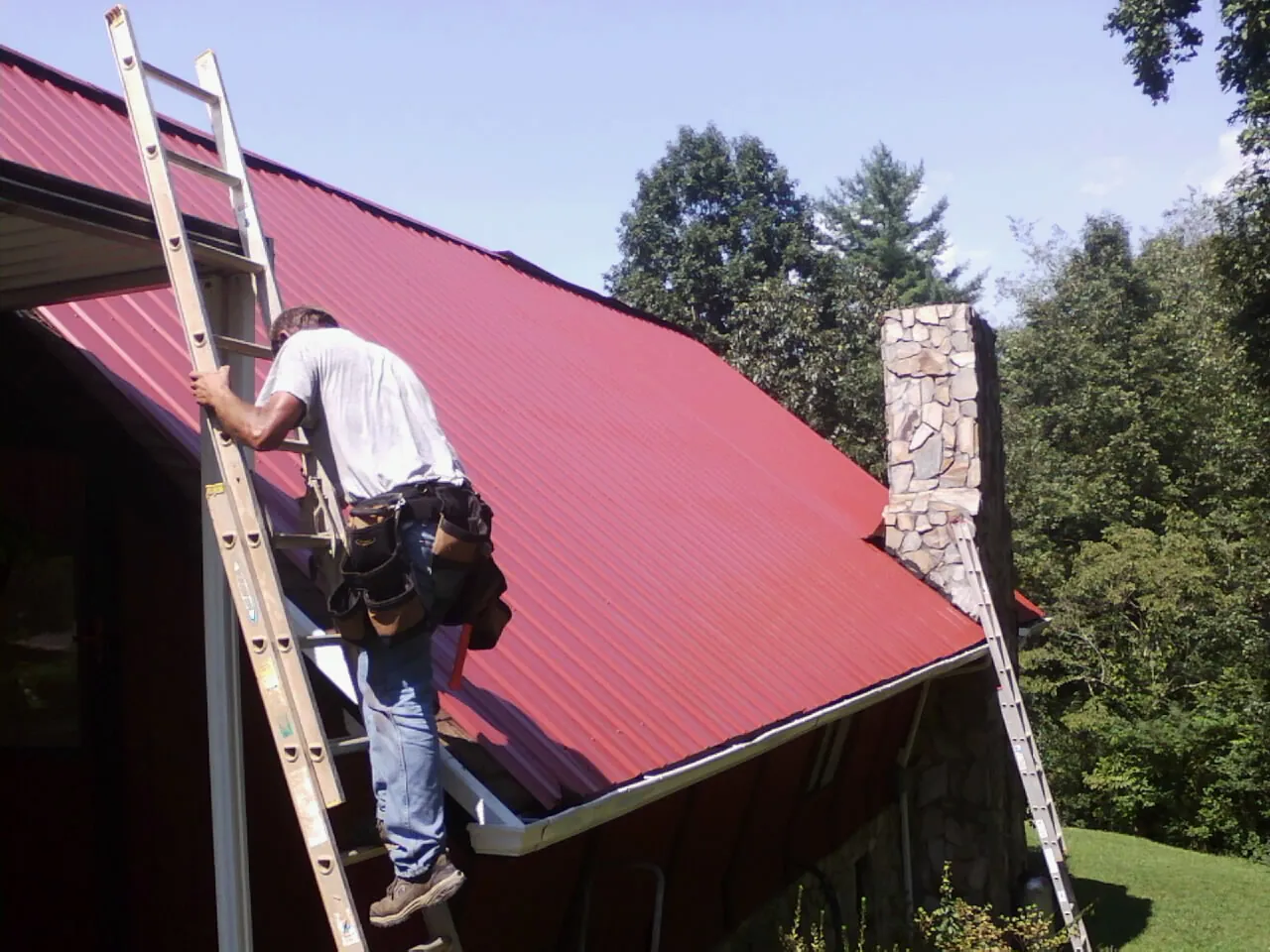 Roofer on a ladder beside a red metal roof and stone chimney — Soffit & Fascia Repair by Roofing Brothers in Angier